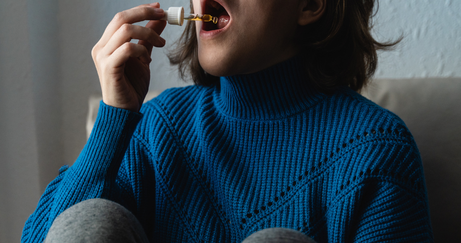 Kobieta aplikuje do ust olejek CBD przez dozownik z fiolki. Ma granatowy sweterek. Widać jej twarz tylko do nosa.