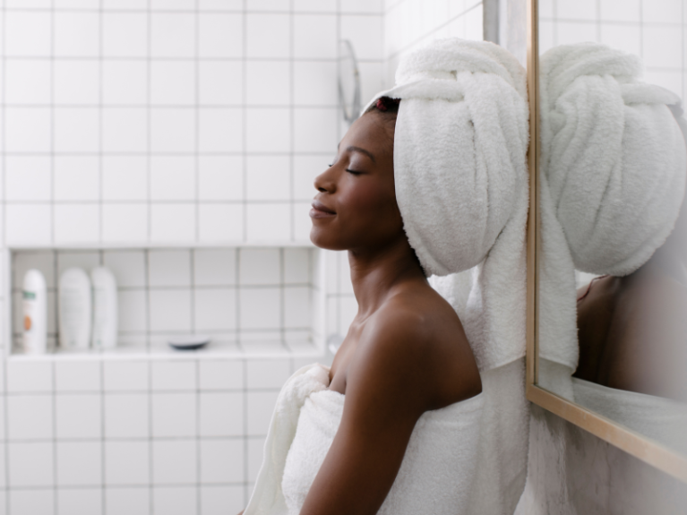 Zrelaksowana kobieta po kąpieli. Stoi w łazience, oparta o lustro. Ma zamknięte oczy. Na jej głowie zawiązany jest turban z ręcznika.